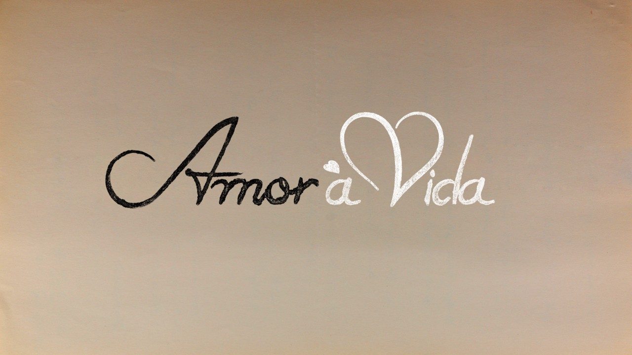 Logotipo da novela Amor à Vida com escrita cursiva, palavra Amor em preto e Vida em branco com um coração estilizado no V, sobre fundo bege claro clara