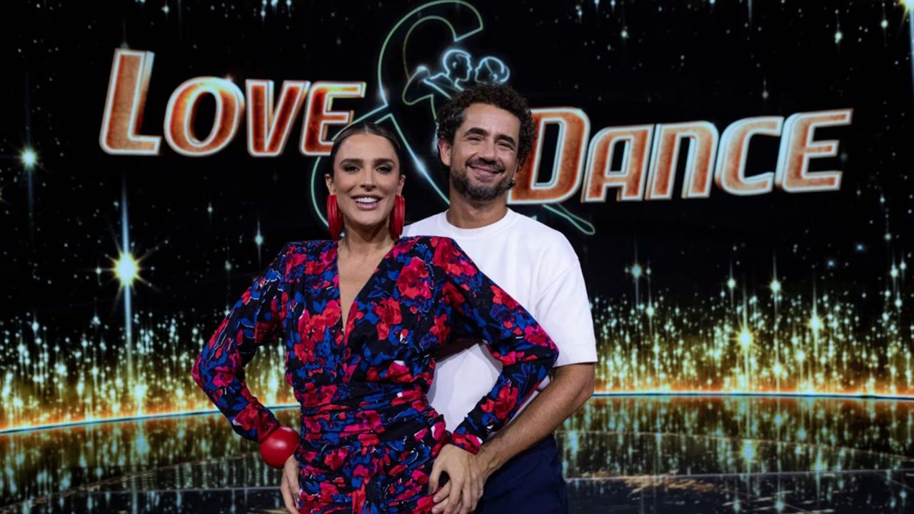 Casal posa sorridente em frente ao letreiro iluminado “Love & Dance”, em um cenário de programa de dança com fundo escuro e luzes douradas. A mulher usa vestido estampado com flores vermelhas e azuis, enquanto o homem veste camiseta branca