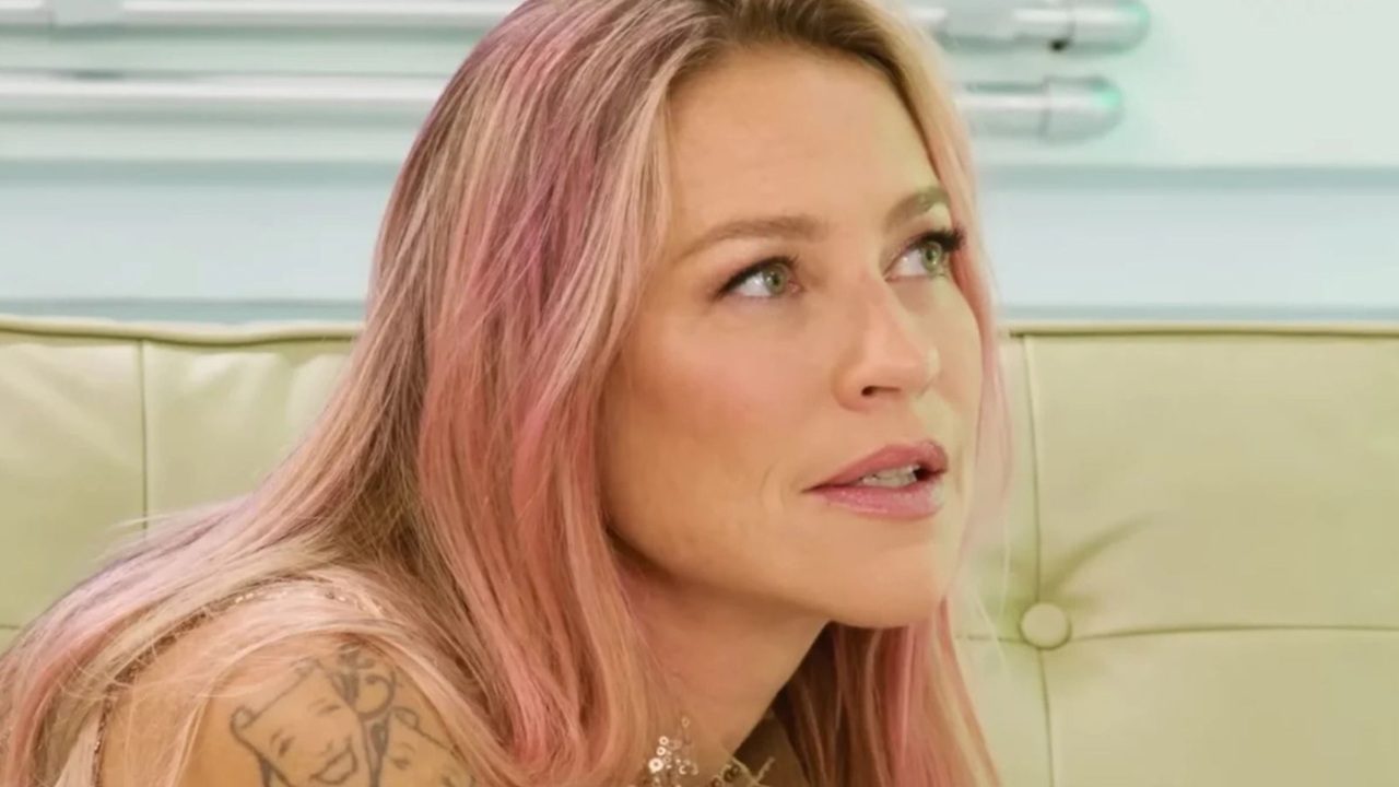 Mulher de cabelos loiros com mechas rosadas olha para o alto com expressão pensativa enquanto está sentada em sofá claro, usando roupa com alças finas e mostrando tatuagem no ombro