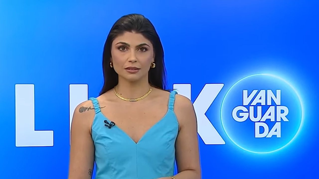 Apresentadora de televisão aparece em estúdio com fundo azul vibrante e o logo “Vanguarda” em destaque. Ela usa vestido azul claro, colar dourado e brincos, além de microfone preso à alça. A jornalista mantém expressão séria enquanto olha diretamente para a câmera, com tatuagens visíveis no ombro e no braço.