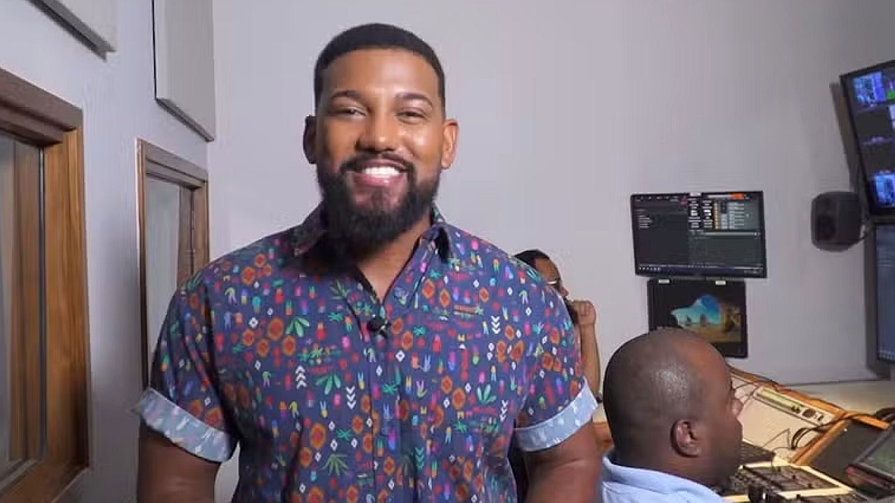 Apresentador sorri nos bastidores de estúdio de TV com equipe técnica e monitores ao fundo