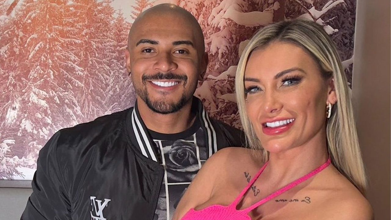 Lucas Ferraz e Andressa Urach posando juntos sorridentes em ambiente interno com quadro de paisagem de neve ao fundo