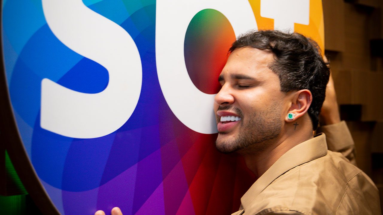 Apresentador Lucas Guimarães de camisa bege, abraçando o logo colorido do SBT de olhos fechados e sorrindo