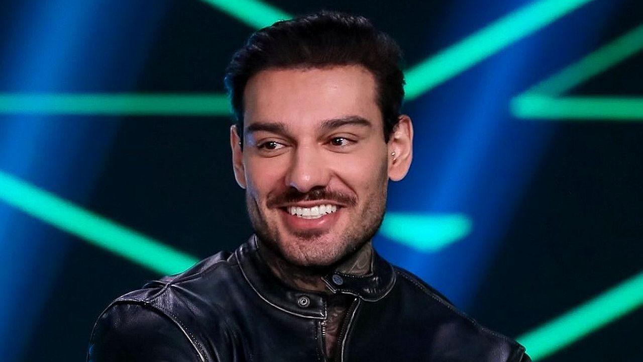 Homem com barba e jaqueta de couro sorri durante participação em programa de TV com luzes neon ao fundo