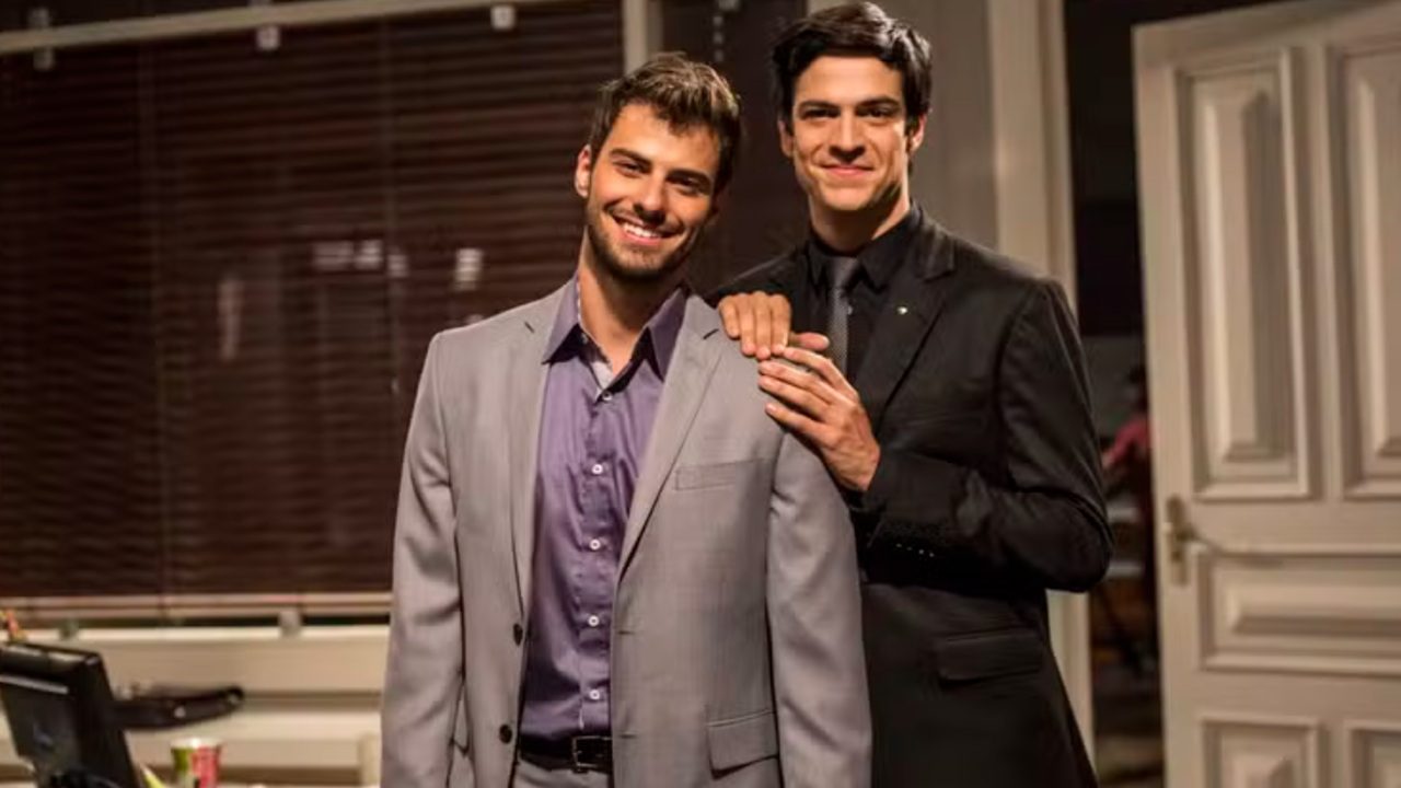 Dois homens posam juntos em um ambiente interno elegante, parecendo um escritório. O homem à frente usa um terno cinza claro com camisa roxa e sorri para a câmera. Atrás dele, outro homem veste um terno preto e apoia as mãos nos ombros do primeiro, também sorrindo. Ao fundo, há persianas escuras e uma porta branca parcialmente aberta, criando um clima formal e acolhedor