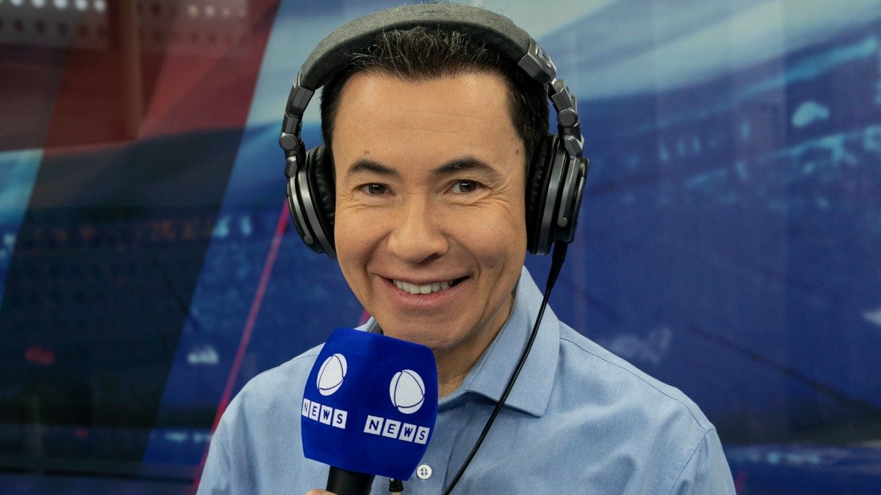 Lucas Pereira sorri usando fones de ouvido e segura microfone com logo da Record News em estúdio de rádio ou TV