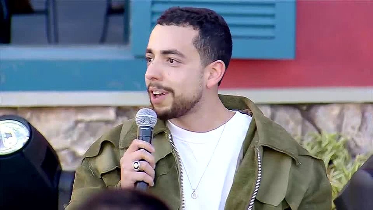 Lucas Selfie fala ao microfone durante participação em programa de TV com jaqueta verde e camiseta branca