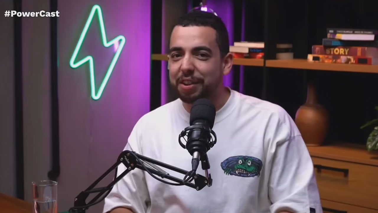 Lucas Selfie fala ao microfone durante gravação do Powercast com cenário iluminado ao fundo