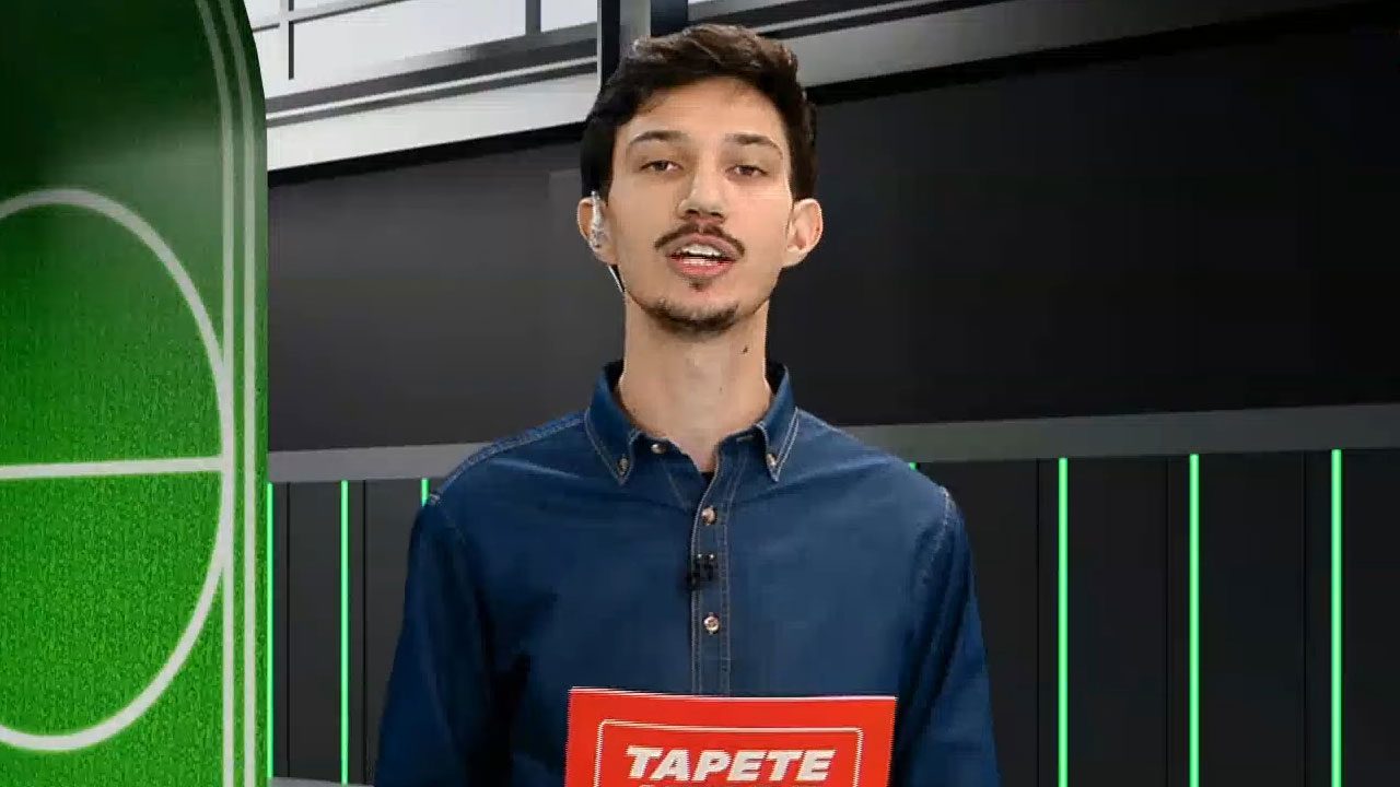 Lucca Dutra, apresentador da Xsports, usando camisa jeans escura e segurando uma ficha vermelha com a palavra Tapete Verde no estúdio