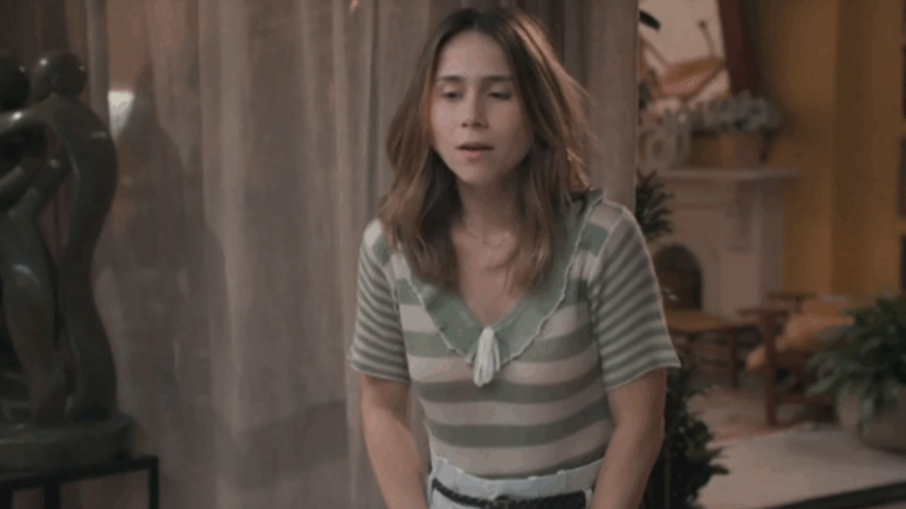 Mulher com expressão de surpresa ou inquietação está em uma sala decorada, vestindo blusa listrada em tons claros. Ao fundo, há cortinas translúcidas, plantas e uma escultura escura, compondo um ambiente acolhedor e elegante