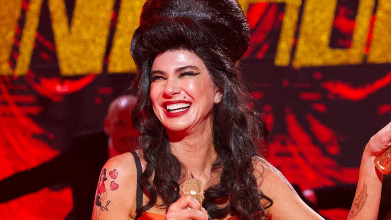 Luciana Gimenez caracterizada como Amy Winehouse sorri no palco do Domingão com Huck segurando microfone dourado