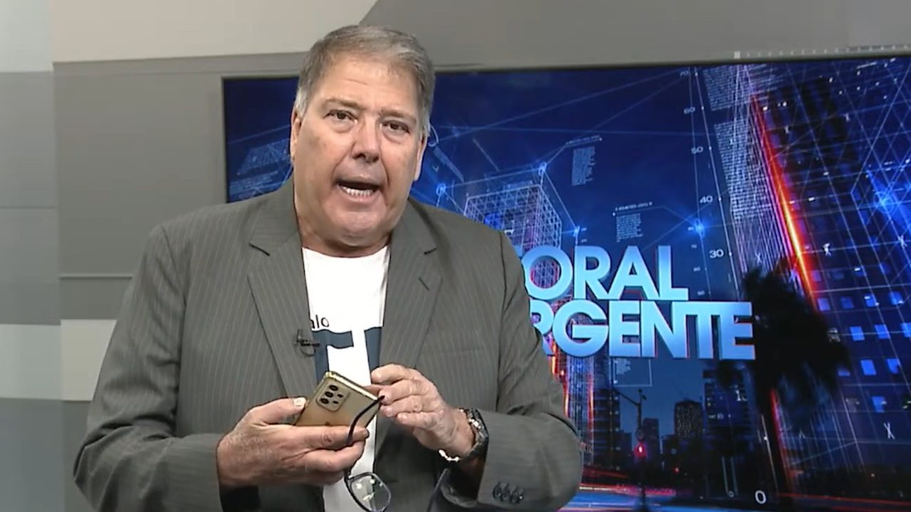 Homem de meia-idade fala diante de um telão com a inscrição “Litoral Urgente”, segurando um celular dourado e um par de óculos na mão. Ele veste blazer cinza sobre camiseta branca, com microfone preso à lapela, em um estúdio de TV com cenário digital de prédios iluminados ao fundo.