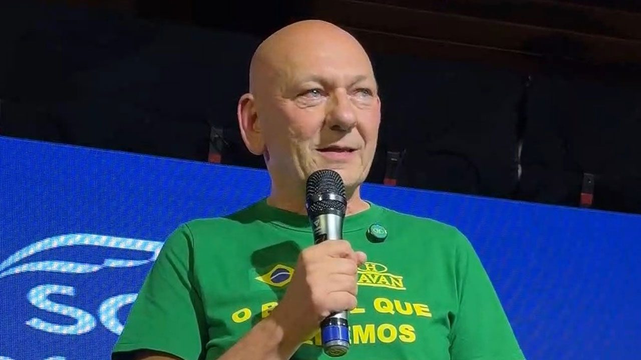 Luciano Hang segura microfone à frente do rosto usando camiseta verde com a bandeira do Brasil e o logotipo da Havan, diante de um painel azul