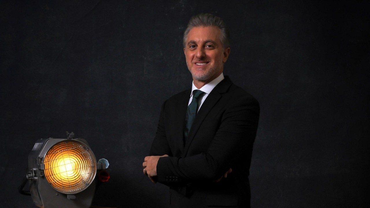 Apresentador Luciano Huck sorri de terno preto e gravata verde escura, posa com braços cruzados ao lado de um refletor de luz aceso em cenário de fundo preto