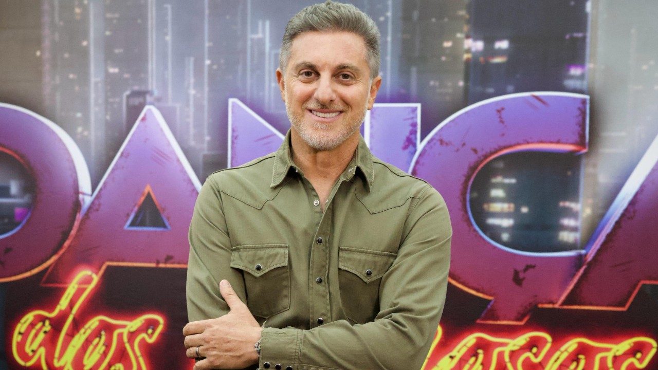 Luciano Huck de camisa verde com braços cruzados em frente a telão com logo da Dança dos Famosos
