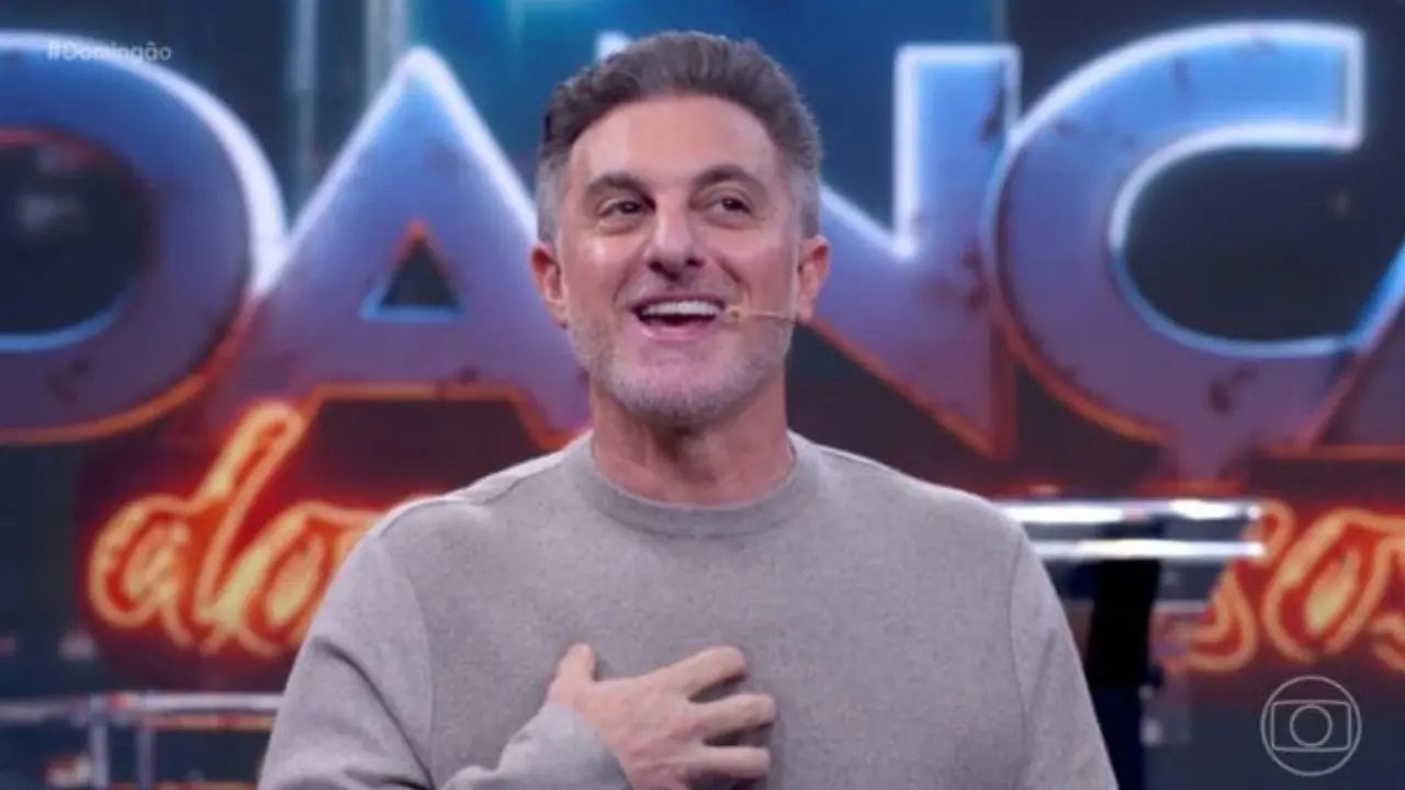 Apresentador sorrindo com microfone de orelha no palco do Domingão em frente a painel iluminado