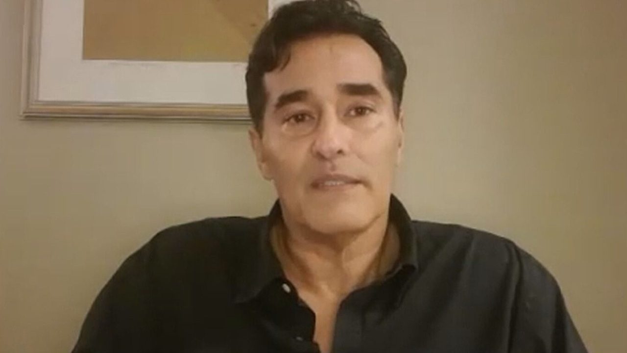 Luciano Szafir com camisa preta fala em vídeo gravado em ambiente interno com parede bege ao fundo