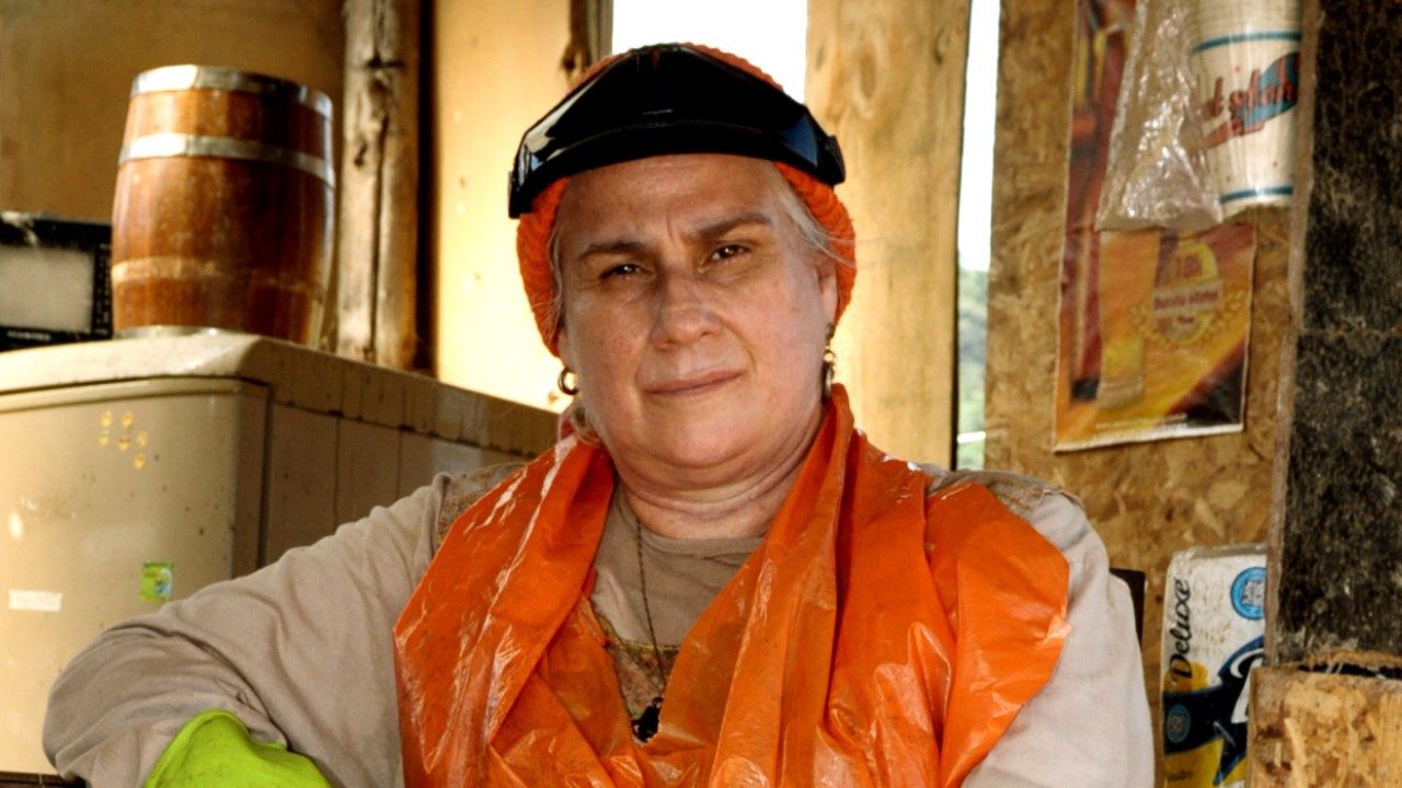 Vera Holtz como Lucinda usa roupas simples e avental laranja com óculos de proteção na cabeça em cenário rústico de Avenida Brasil
