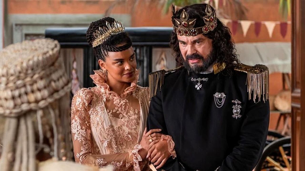 Homem e mulher com coroas e trajes reais conversam em cena de época com figurinos luxuosos