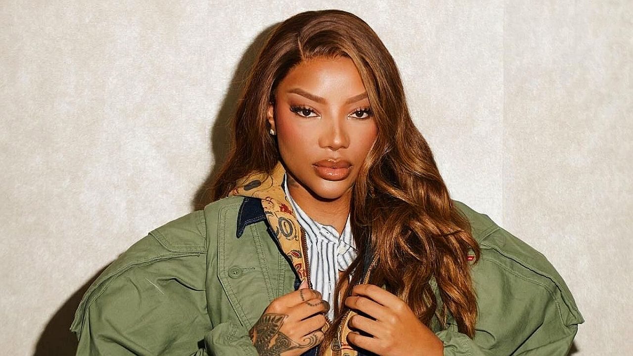 Mulher posa com jaqueta verde e cabelo longo ondulado em ensaio fotográfico contra fundo neutro