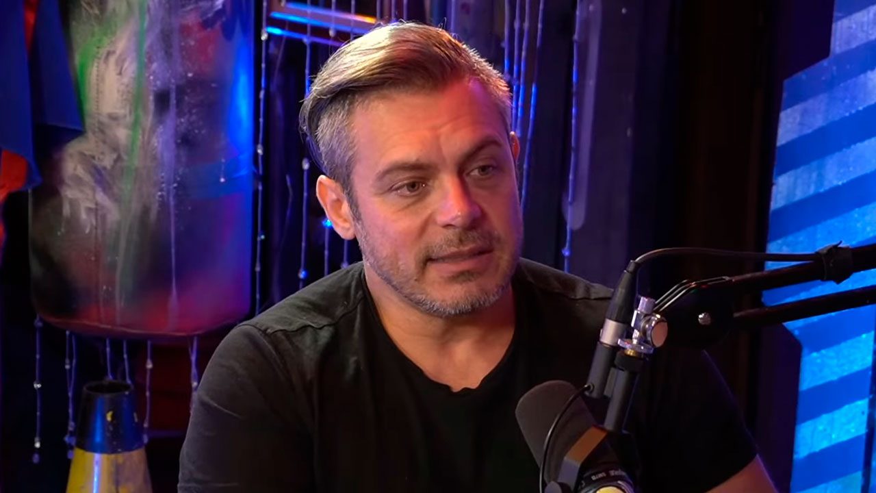 Luigi Baricelli em entrevista no estúdio de um podcast, usando camiseta preta e falando ao microfone
