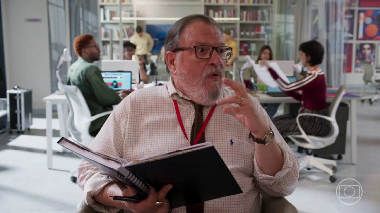 Luis Melo caracterizado como chefe de redação em cena ambientada em escritório moderno