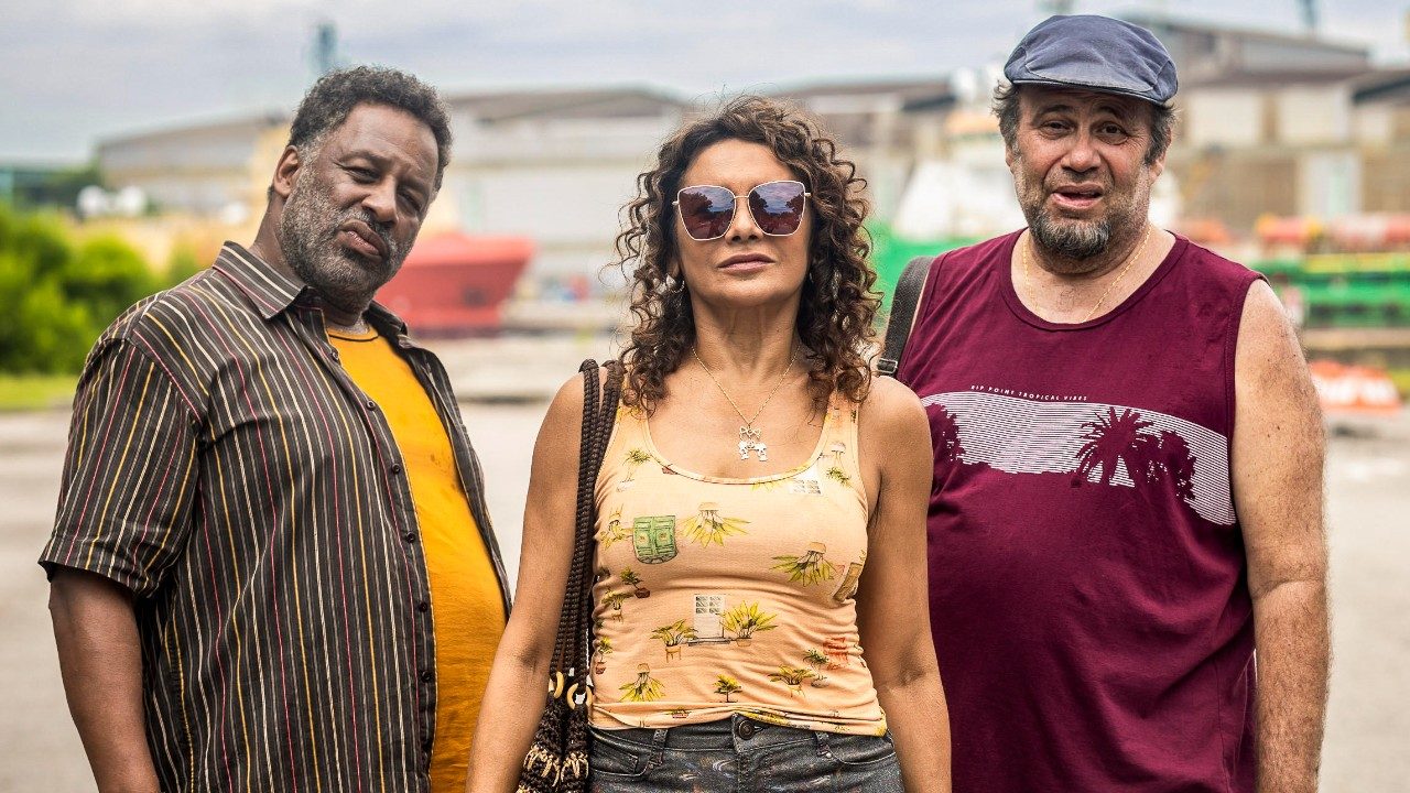 Ailton Graça, Dira Paes e Otávio Muller posam como Luisão, Conceição e Pablo, Dira usa óculos escuros e regata bege, Otávio usa boina e regata bordô