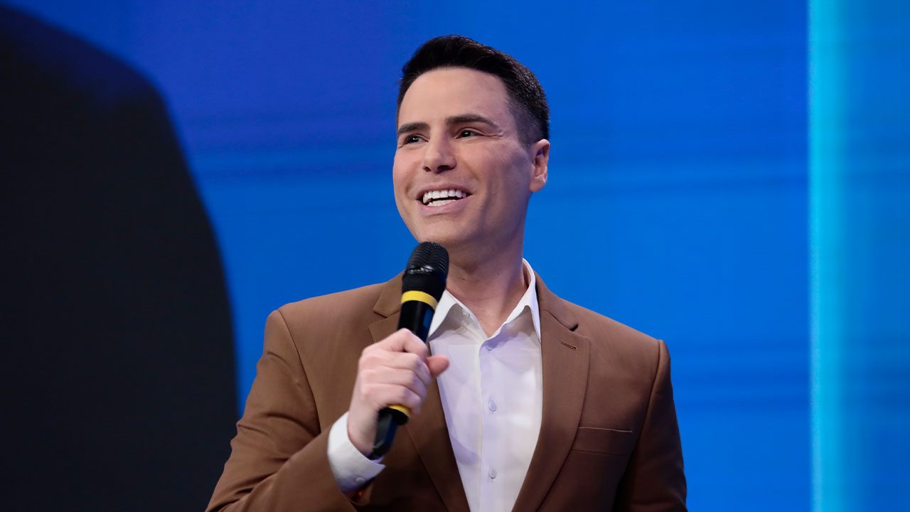 Luiz Bacci segurando microfone durante coletiva da nova programação do SBT em cenário azul