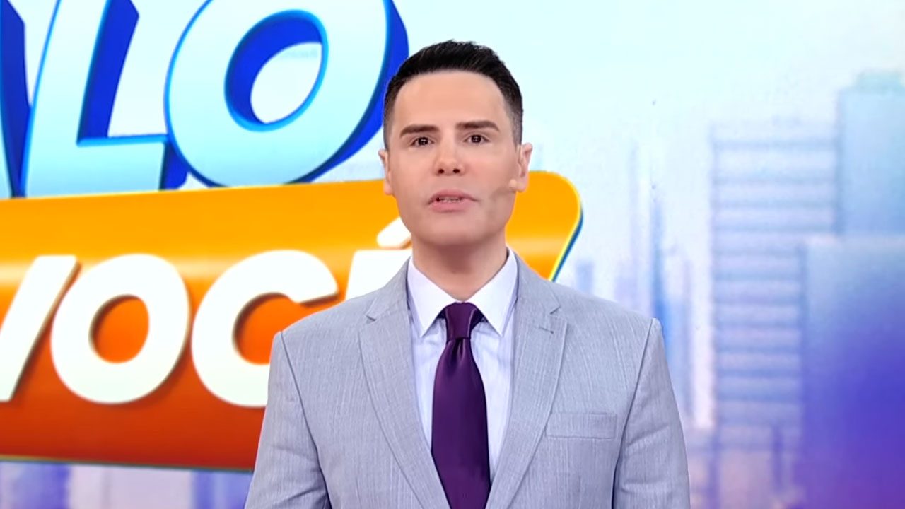 Luiz Bacci apresenta o Alô Você no SBT vestindo terno cinza e gravata roxa diante do logotipo do programa