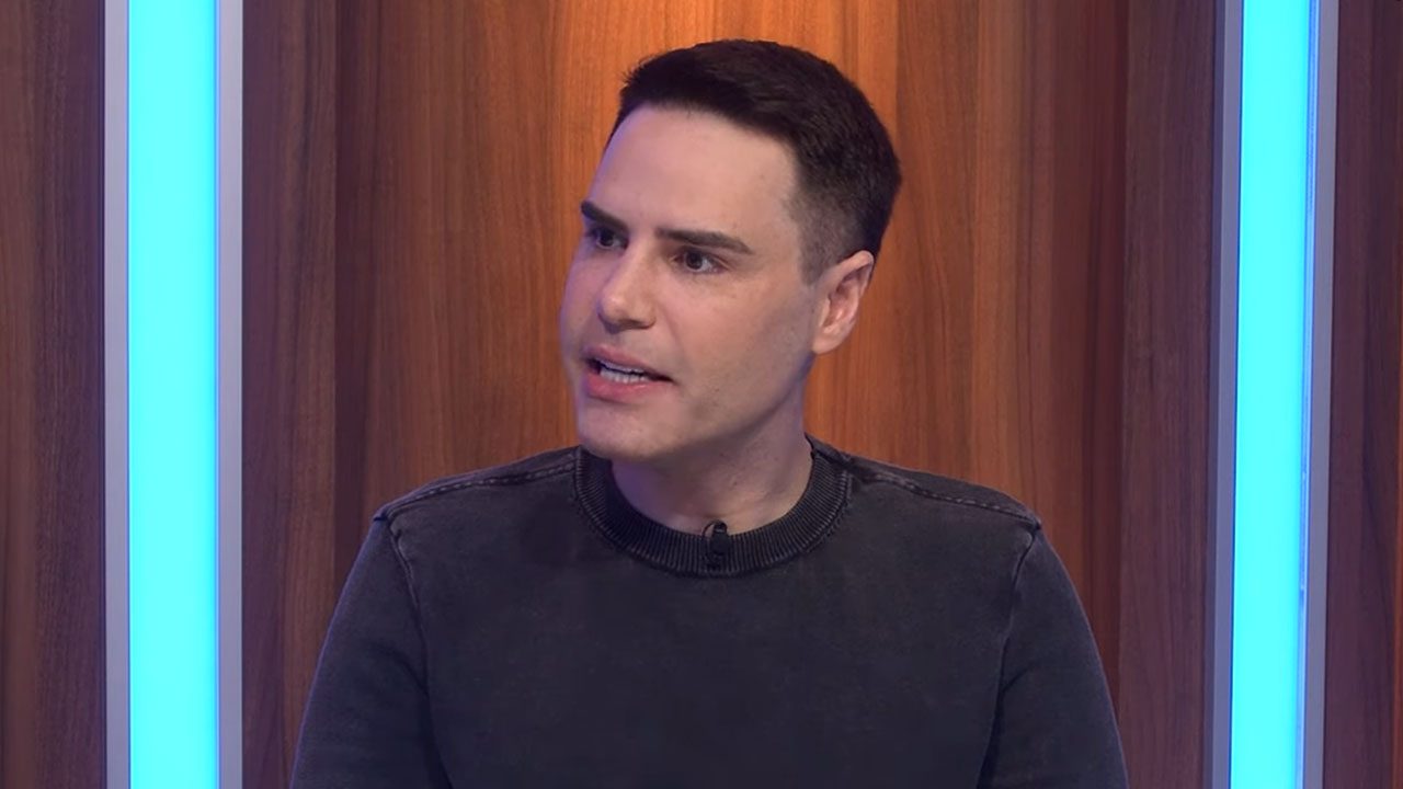 Luiz Bacci falando durante participação no Programa Flávio Ricco diante de painel com luzes azuis