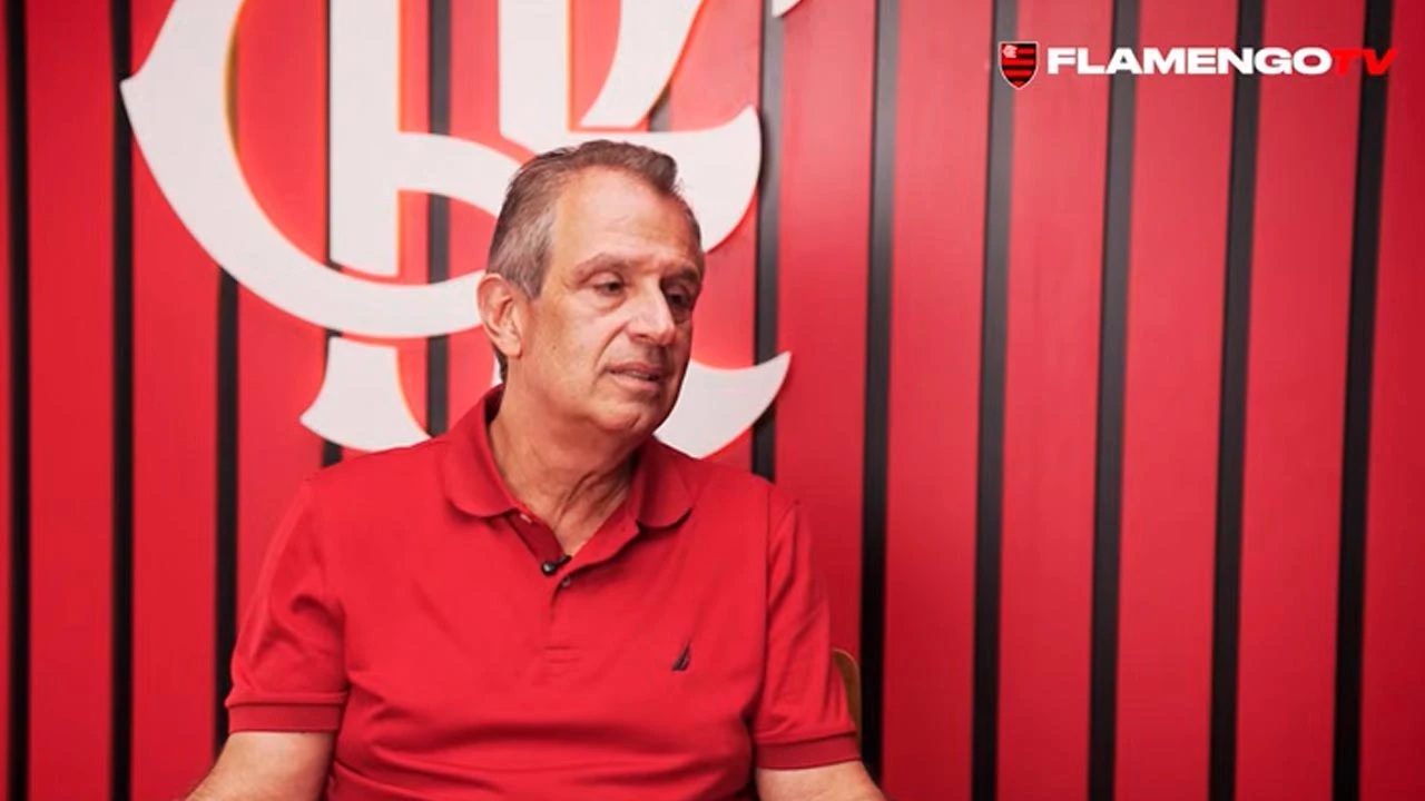Homem adulto sentado em ambiente interno com fundo institucional do Flamengo, pele clara cabelos curtos grisalhos penteados para trás expressão séria e concentrada olhando levemente para o lado esquerdo da imagem, veste camisa polo vermelha de manga curta com pequeno logotipo no peito, segura as mãos próximas ao corpo em posição de conversa ou entrevista, atrás dele parede vermelha com listras verticais pretas e grande símbolo estilizado do Flamengo em branco parcialmente visível, no canto superior direito aparece a marca Flamengo TV, iluminação uniforme e frontal típica de estúdio ou sala de entrevistas transmite contexto de fala oficial ou depoimento relacionado ao clube