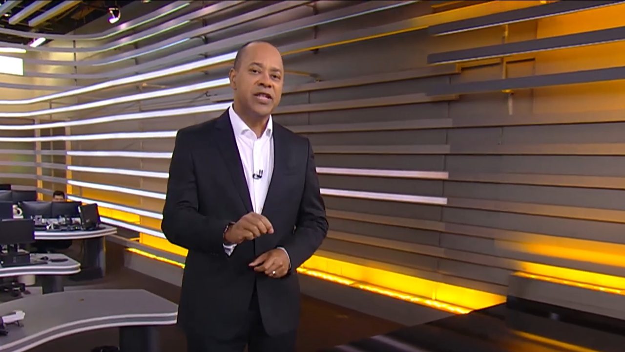 Apresentador de terno escuro e camisa branca fala em estúdio de telejornal com fundo moderno iluminado em tons de amarelo e branco, com mesas e computadores ao fundo