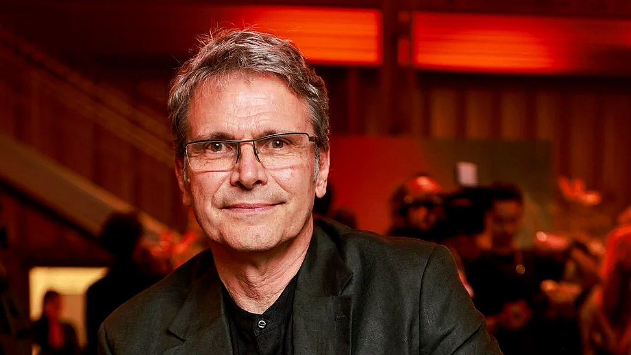 Homem de óculos e terno preto posa em evento com iluminação vermelha ao fundo