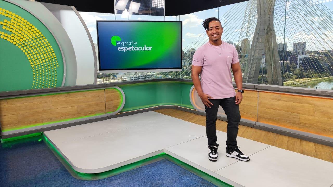 Homem adulto de pele morena está em pé em um estúdio de televisão moderno usando camiseta rosa clara de manga curta calça preta ajustada e tênis pretos com detalhes brancos ele sorri levemente para a câmera com postura relaxada braços soltos ao lado do corpo e cabelo curto com tranças ou dreads presos no topo do lado esquerdo ao fundo há um cenário predominantemente verde com painéis curvos detalhes em madeira clara e iluminação embutida em LED verde um grande monitor à esquerda exibe o texto esporte espetacular em letras brancas sobre fundo verde com um símbolo circular no canto esquerdo