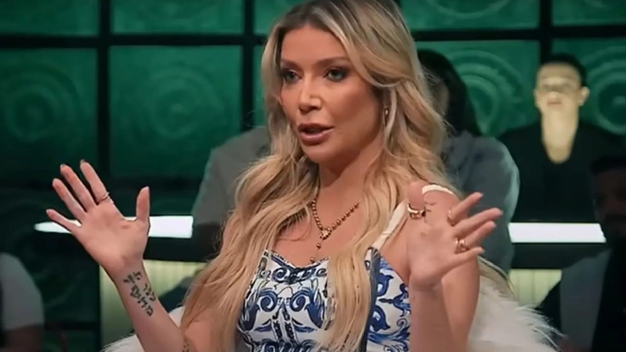 A imagem mostra uma mulher loira de cabelos longos, vestindo um vestido branco e azul estampado, com colar dourado e anéis. Ela está sentada em um estúdio de TV, com as mãos levantadas enquanto fala, aparentando explicar ou enfatizar algo