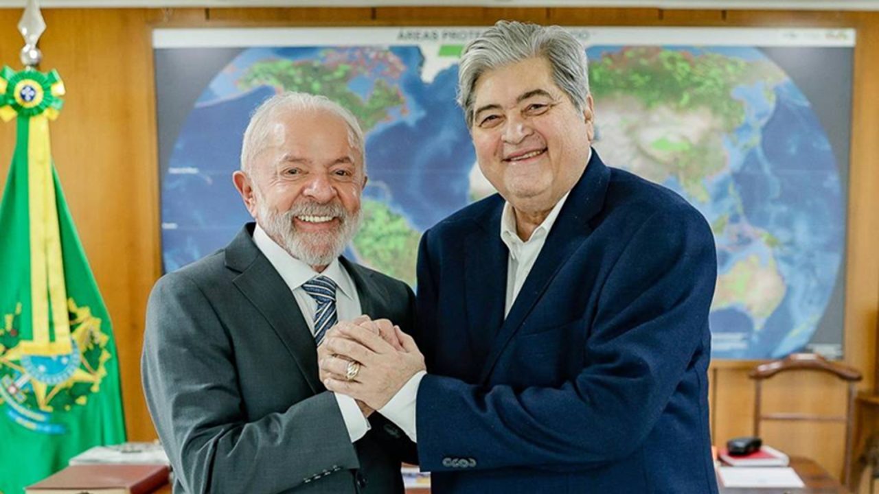 A imagem mostra Lula e Datena sorridentes dando um aperto de mãos em um ambiente formal, com uma bandeira do Brasil e um mapa-múndi ao fundo. Ambos vestem ternos escuros e aparentam estar em um gabinete ou escritório oficial, em clima cordial e amistoso