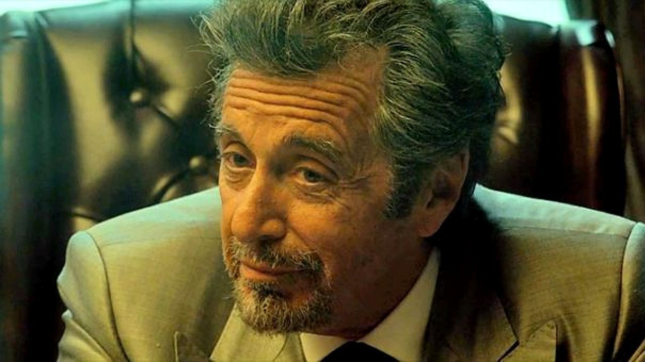 Homem grisalho de terno inclina a cabeça e observa com expressão pensativa em ambiente interno sofisticado