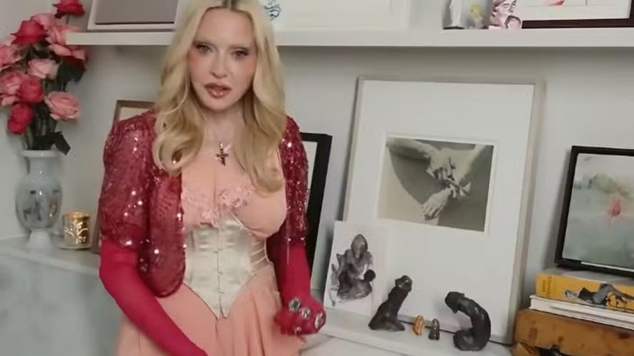 Madonna usa vestido rosa com corpete, bolero vermelho de paetês e luvas enquanto posa diante de estante com quadros, flores e esculturas em ambiente interno