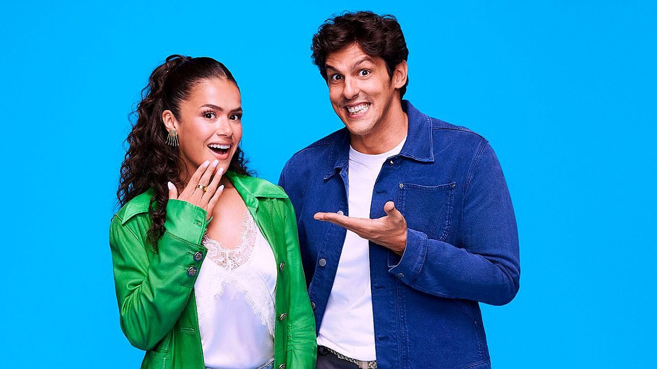 Dupla de apresentadores sorri para foto promocional com fundo azul em campanha de programa de entretenimento