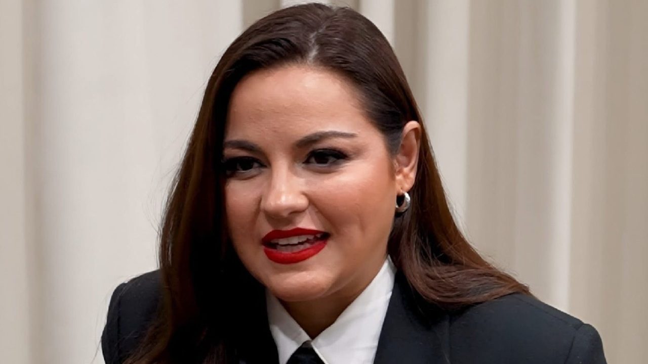 A imagem mostra uma mulher de cabelos castanhos lisos e maquiagem marcante, com batom vermelho e delineador preto. Ela está vestindo um terno preto com camisa branca e gravata, transmitindo um visual elegante e formal. O fundo é neutro, com cortinas claras