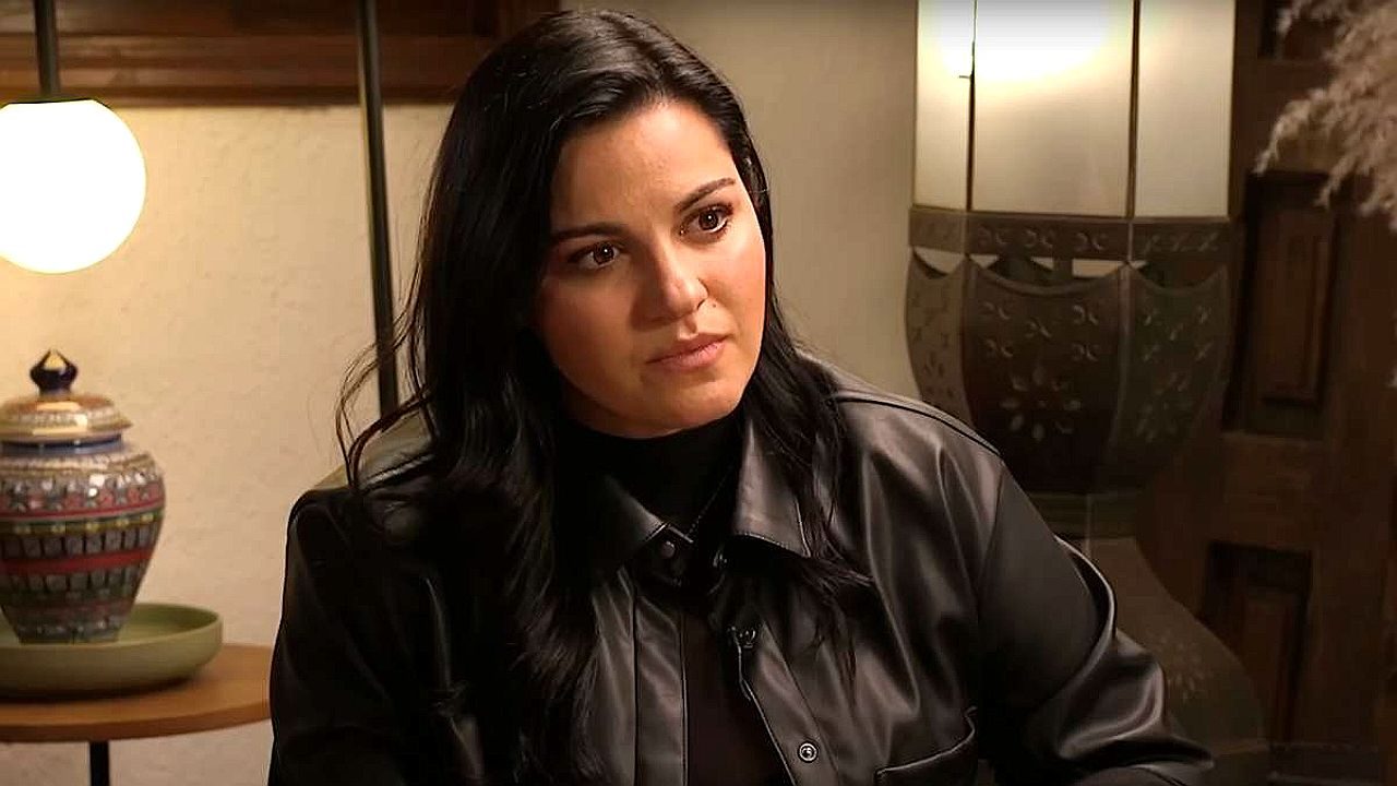 Mulher de cabelos escuros observa alguém com expressão séria durante conversa em ambiente interno iluminado, com luminária e objetos decorativos ao fundo