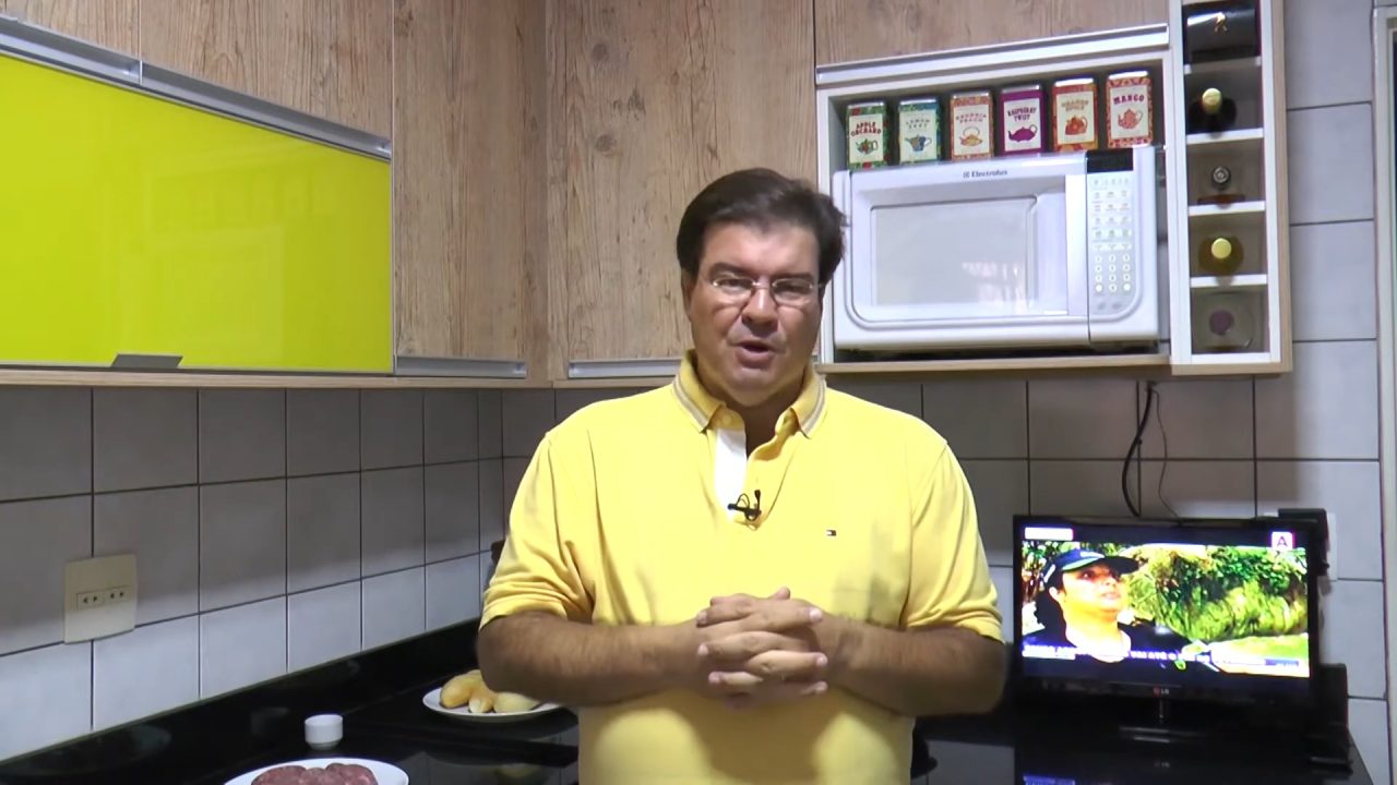 Manoel Branco, vestindo uma camisa amarela, fala para a câmera dentro do estúdio de seu programa na TV Clube, afiliada da Band. Ao fundo, há armários de madeira, um micro-ondas embutido com caixas de mantimentos acima e uma pequena televisão ligada sobre a bancada.