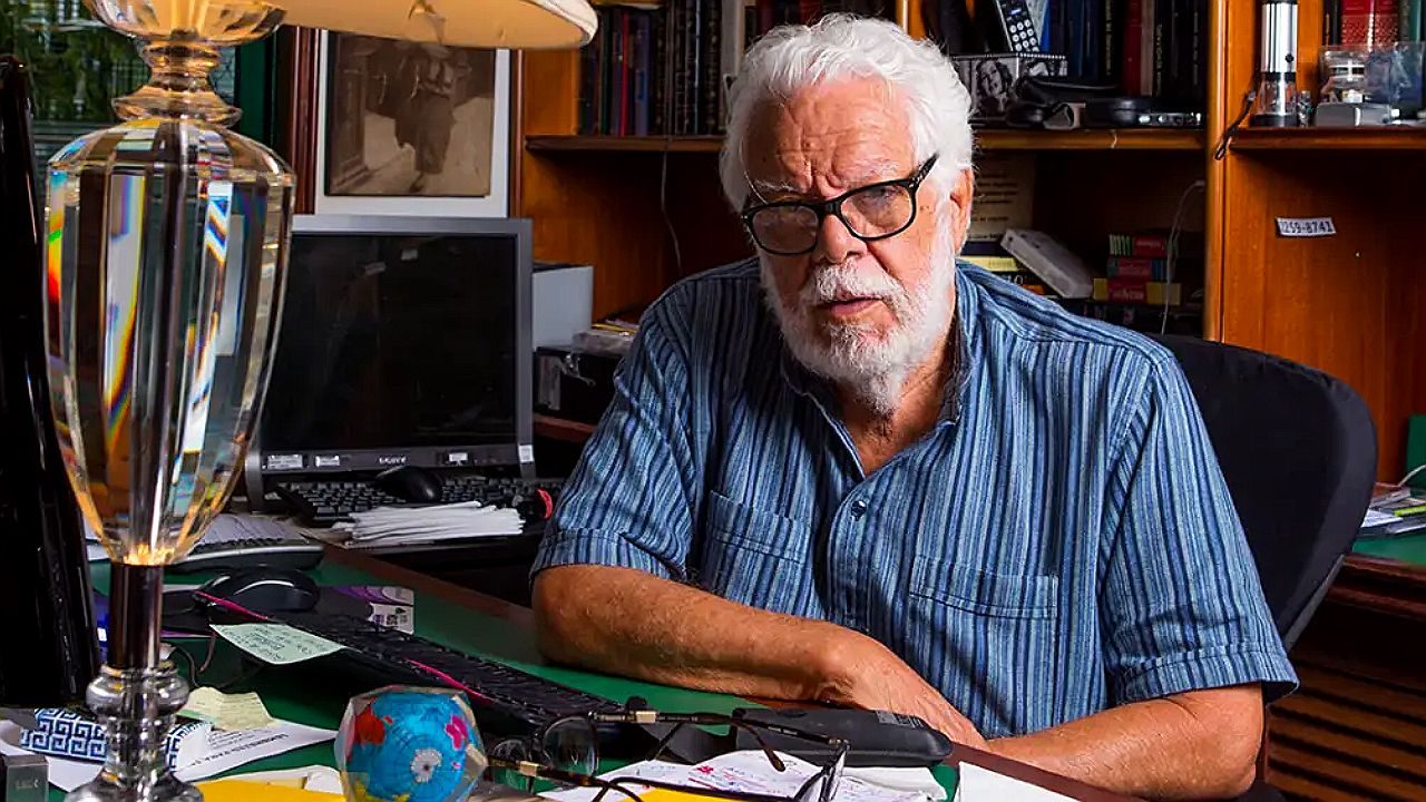 Homem idoso de óculos e camisa listrada sentado em escritório com computador e papéis sobre a mesa