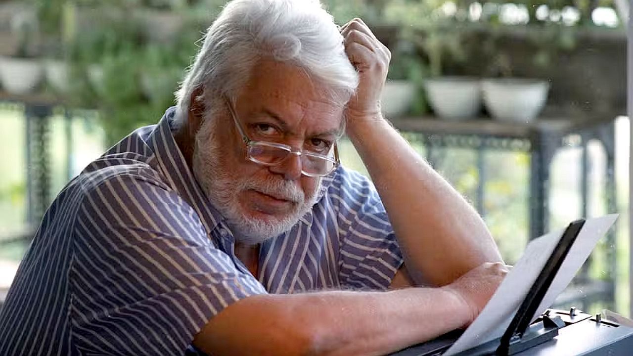 Homem grisalho com óculos lê documento apoiado em mesa enquanto observa atento em ambiente residencial