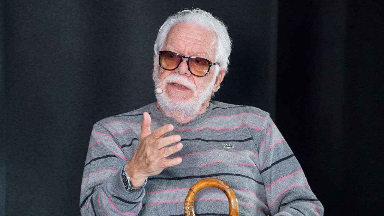 Manoel Carlos usa óculos escuros e suéter listrado cinza enquanto fala com microfone preso ao rosto segurando uma bengala