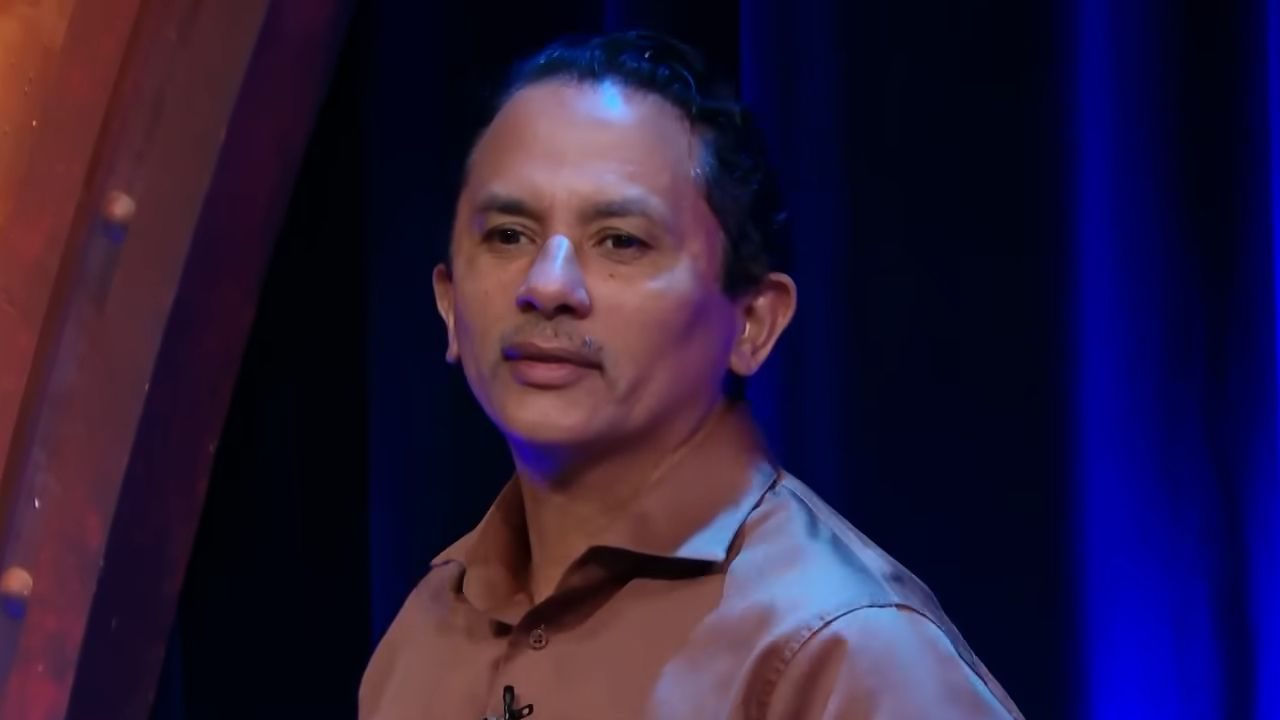 Cantor Manoel Gomes veste camisa social marrom e encara a câmera seriamente, posando em estúdio com iluminação azul e fundo escuro com cortinas