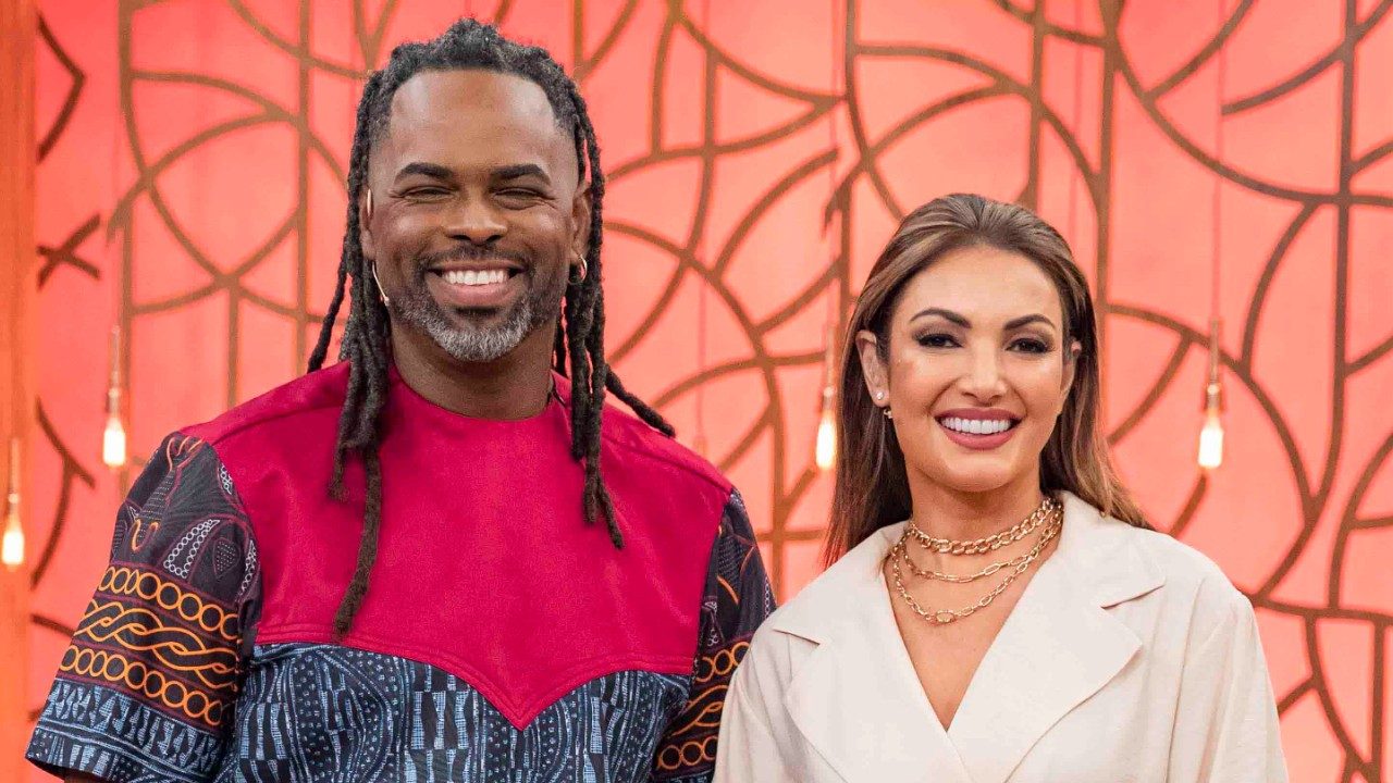 Manoel Soares e Patrícia Poeta sorrindo lado a lado em estúdio com fundo laranja decorado com luzes pendentes