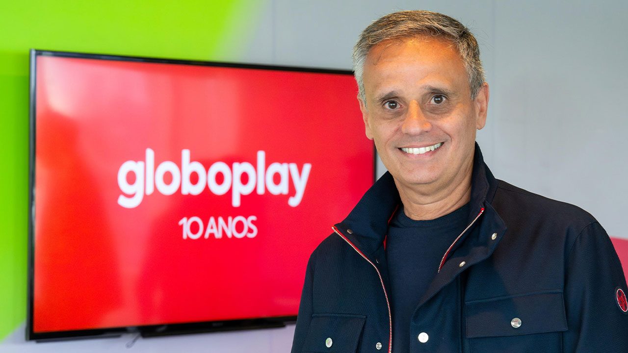O executivo Manuel Belmar sorri usando jaqueta escura e camisa polo preta em frente a um telão com o logo Globoplay 10 anos