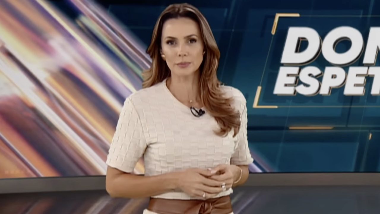 Manuela Queiroz, de blusa clara, apresenta o Domingo Espetacular em estúdio com painel gráfico ao fundo