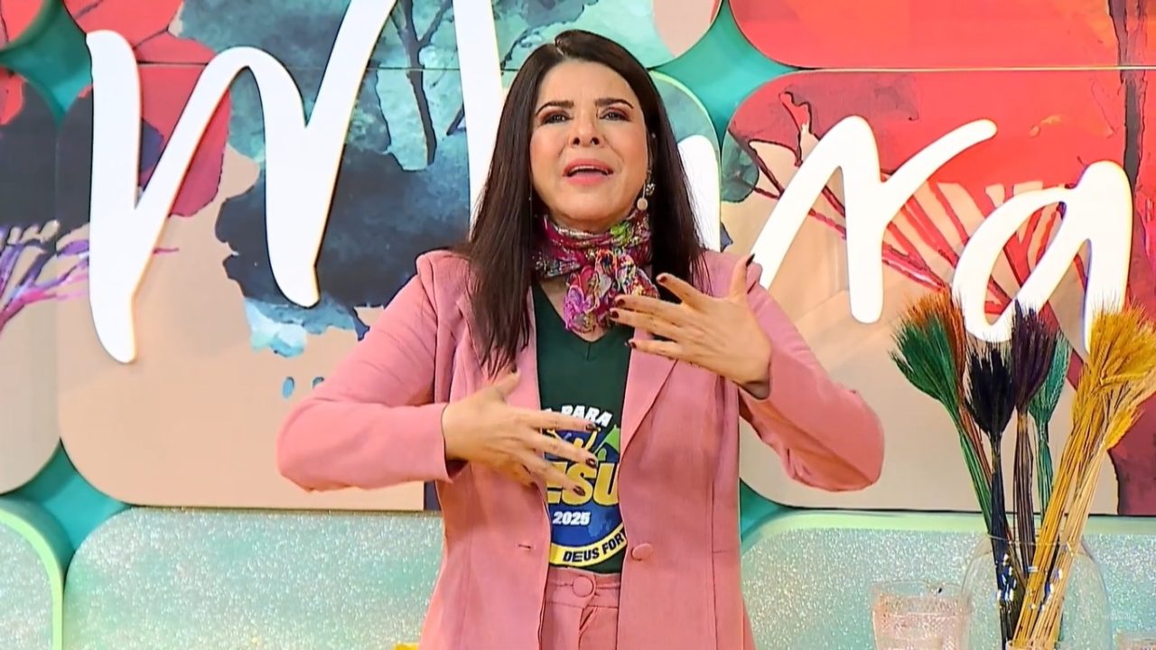 Mara Maravilha falando no palco do programa Mulheres, com figurino rosa e cenário colorido ao fundo